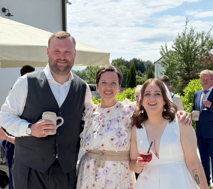 Drei lächelnde Personen draußen, ein Mann mit einer Kaffeetasse und zwei Frauen, eine mit einem Glas Rotwein.