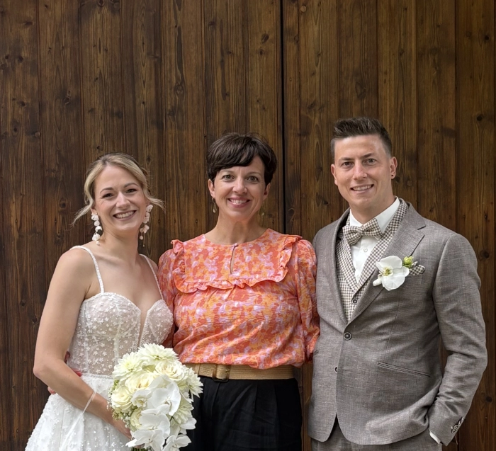 Drei lächelnde Personen vor einer Holzwand, links eine Braut im weißen Kleid mit Blumenstrauß, rechts ein Bräutigam im grauen Anzug, in der Mitte eine Frau mit orange gemusterter Bluse.