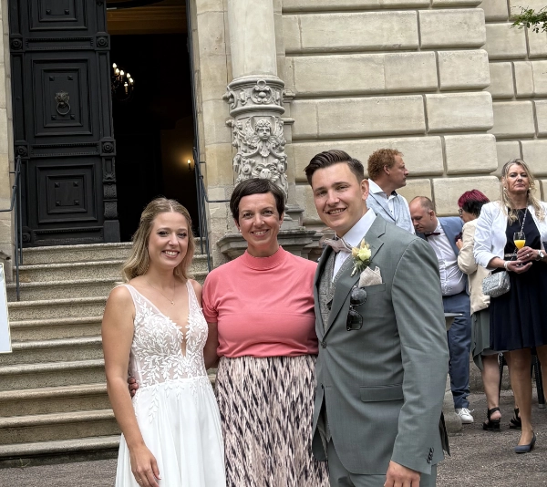 Eine Braut in einem weißen Spitzenkleid, eine Frau in einem rosa Oberteil und gemustertem Rock sowie ein Bräutigam im grauen Anzug posieren lächelnd vor einer Treppe mit historischer Gebäudefassade.
