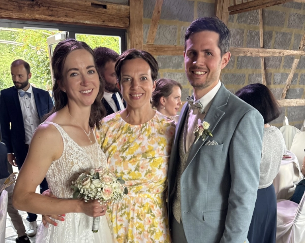 Braut in weißem Spitzenkleid mit Blumenstrauß steht links neben Frau in gelb-weiß gemustertem Kleid und Bräutigam im grauen Anzug mit Fliege in einer Veranstaltungshalle mit Holzbalkenwand.