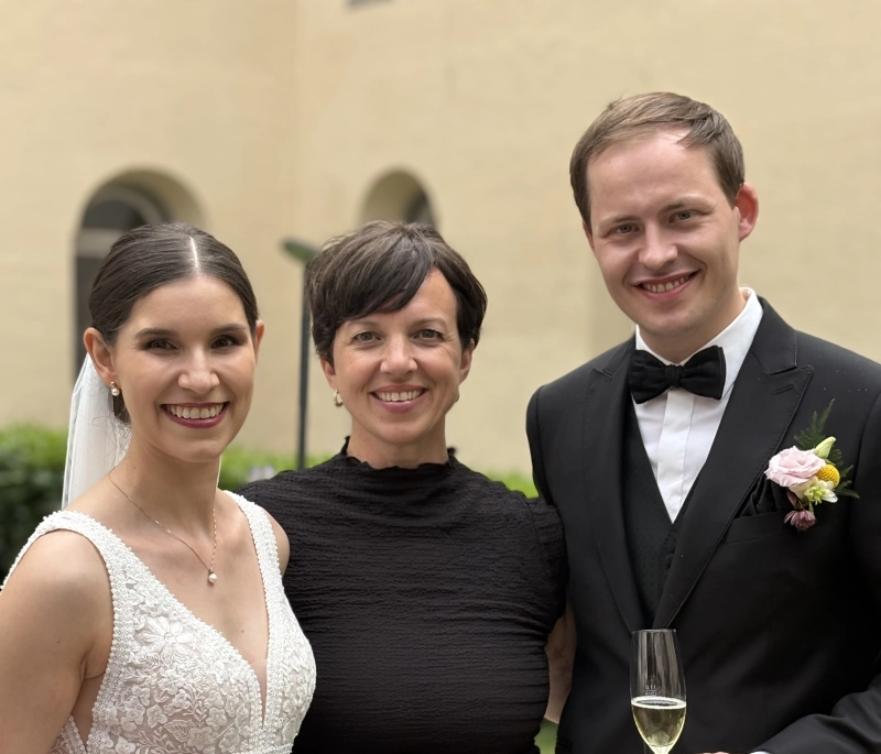 Braut in weißem Spitzenkleid, Frau in schwarzem Kleid und Bräutigam im schwarzen Anzug mit Fliege und Blumenanstecker, die lächelnd in einem Innenhof posieren.