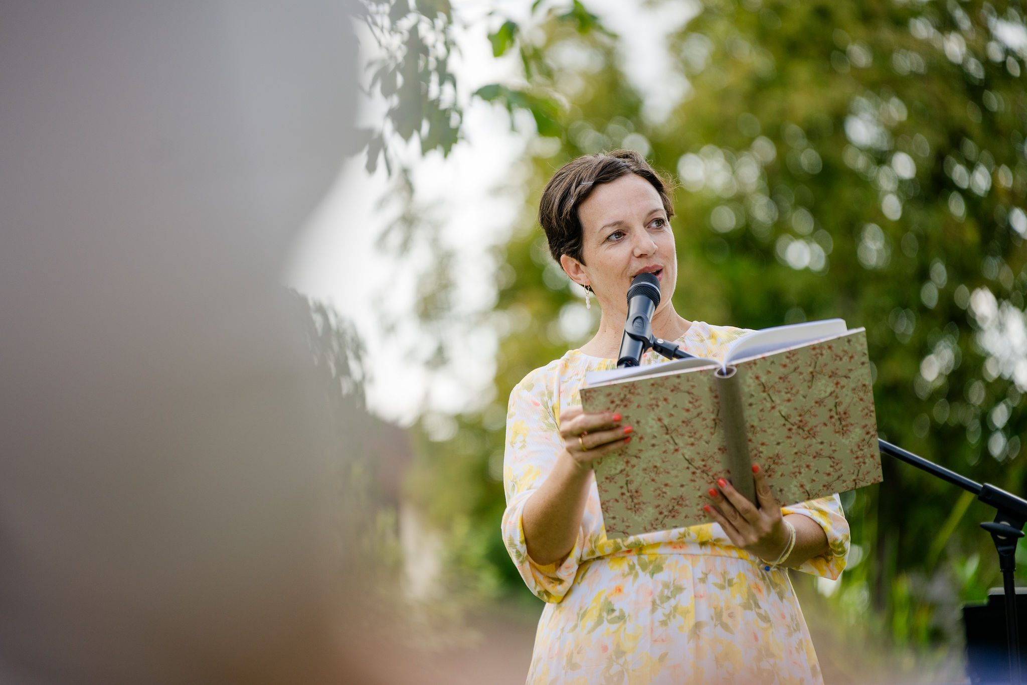 Anna Fasciani - freie Traurednerin -  liest aus einem Buch in einem blumigen Kleid vor einem Mikrofon im Freien.