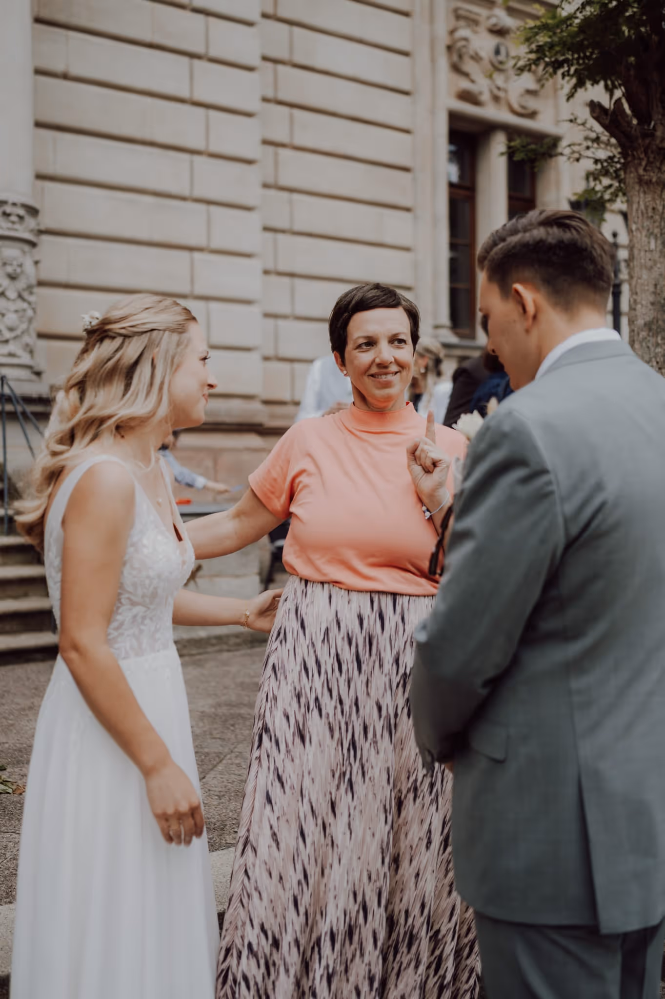 Brautpaar im Gespräch mit Anna Fasciani - freie Traurednerin - in einem gemusterten Rock und rosa Oberteil vor einem historischen Gebäude.