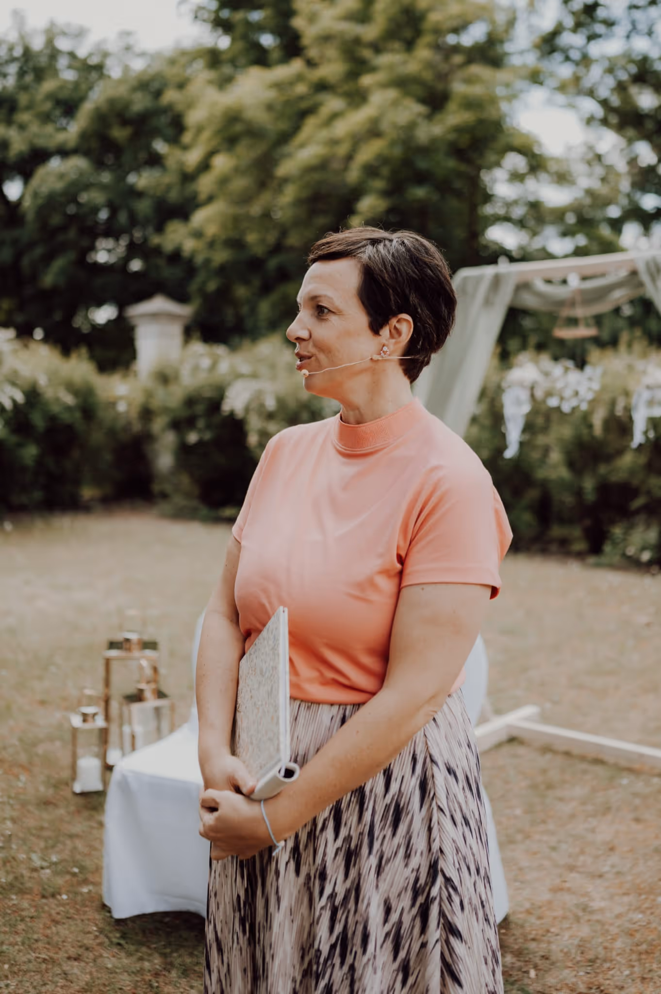 Anna Fasciani - freie Traurednerin -  mit kurzem Haar und Mikrofonsystem hält ein Buch und steht in einem Garten mit dekorativer Kulisse.