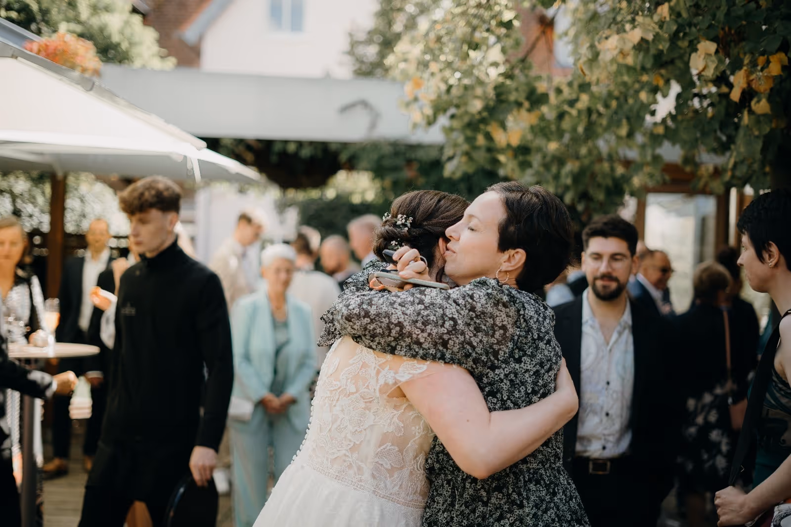 Zwei Frauen umarmen sich herzlich bei einer Feier im Freien unter Bäumen, umgeben von Gästen.
