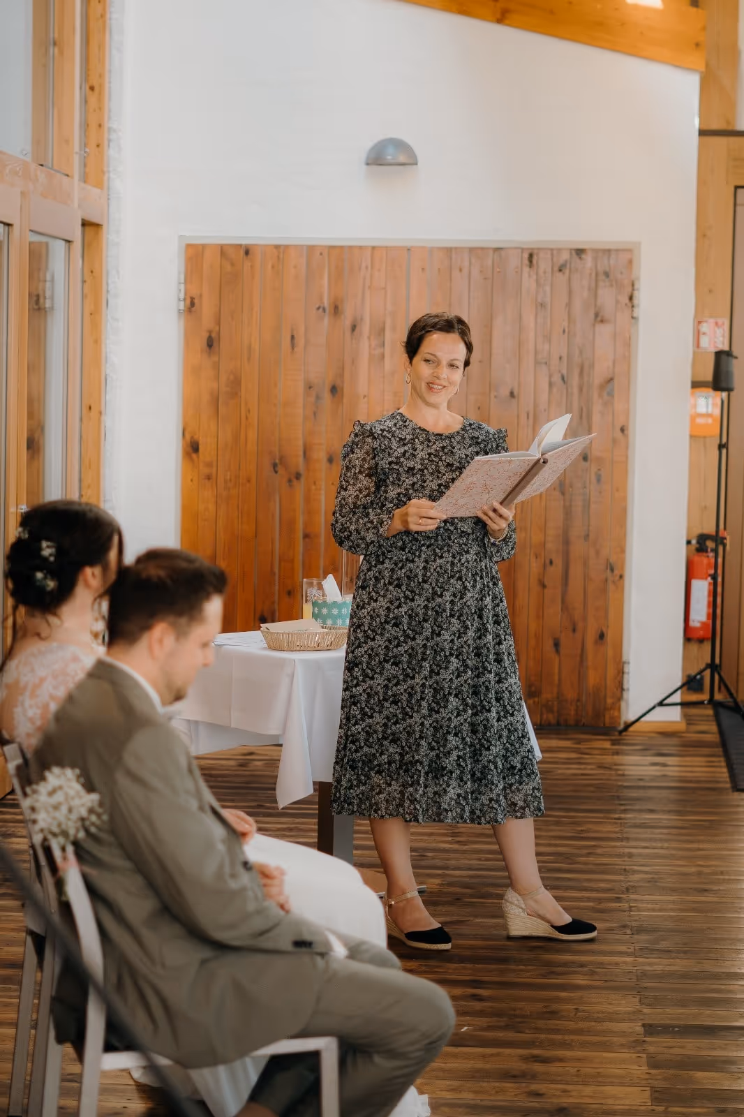 Anna Fasciani - freie Traurednerin -  in einem schwarzen Blumenkleid liest aus einem Buch, während ein Brautpaar ihr aufmerksam zuhört, sitzend auf Holzstühlen in einem saalähnlichen Raum mit Holzboden und Holzwand.