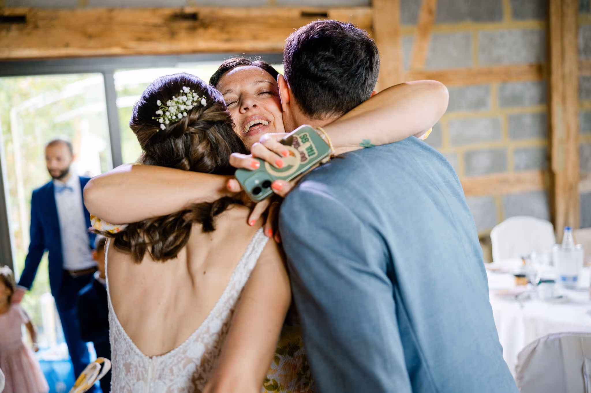 Drei Menschen umarmen sich herzlich bei einer Hochzeit, weibliche Braut mit Blumen im Haar, glückliche Gesichter.