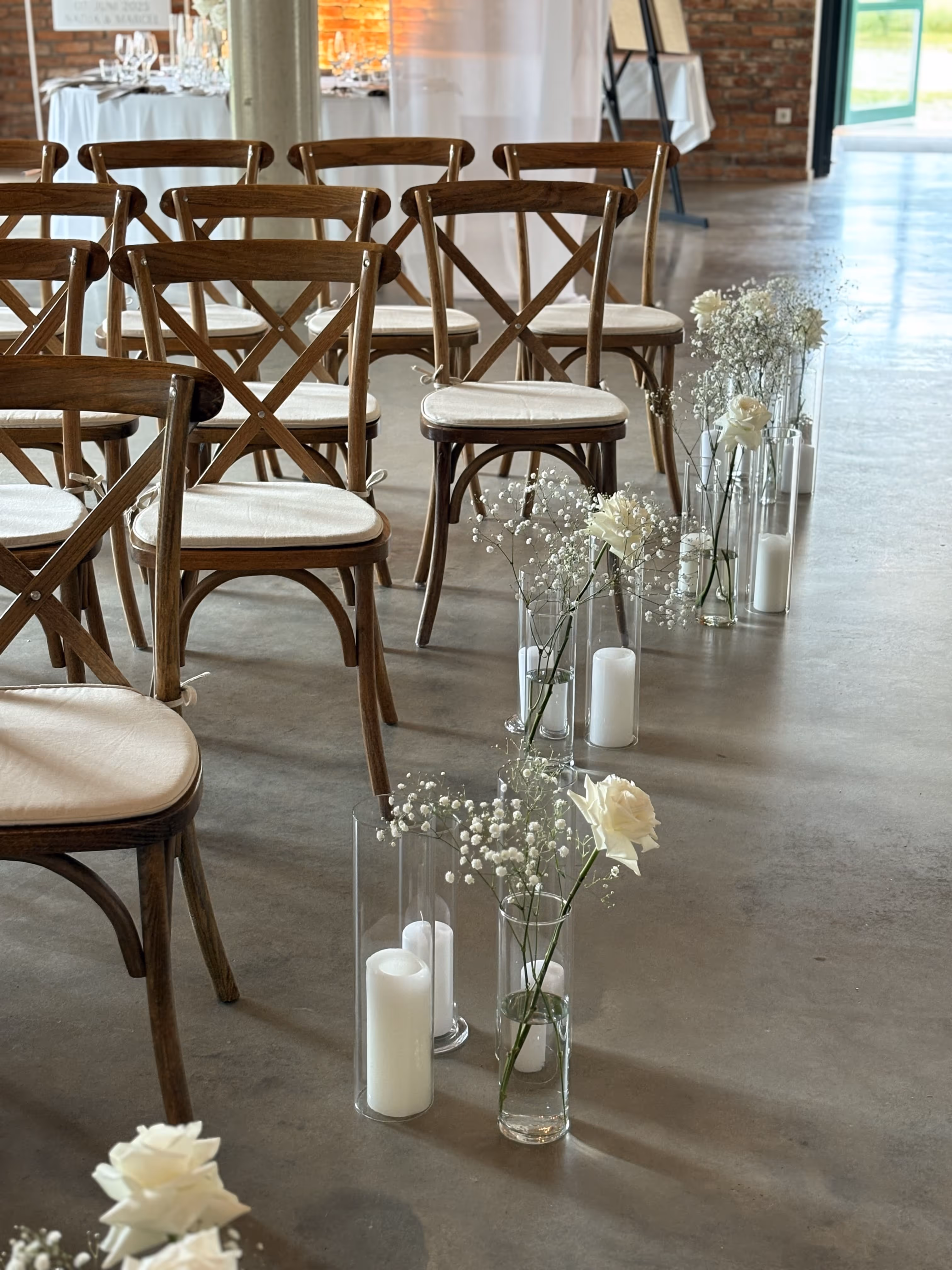 Reihen aus braunen Holzstühlen mit weißen Sitzkissen, entlang eines Gangs dekoriert mit weißen Kerzen und zarten weißen Blumen in Glasvasen.