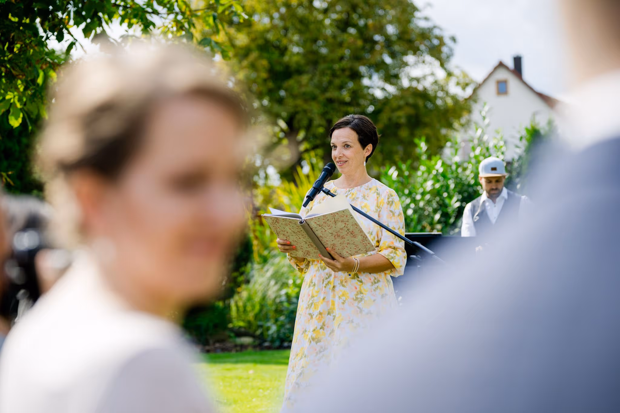 Anna Fasciani - freie Traurednerin -  liest bei freier Trauung im Garten aus einem Buch mit Mikrofon, im Hintergrund Mann mit Basecap.