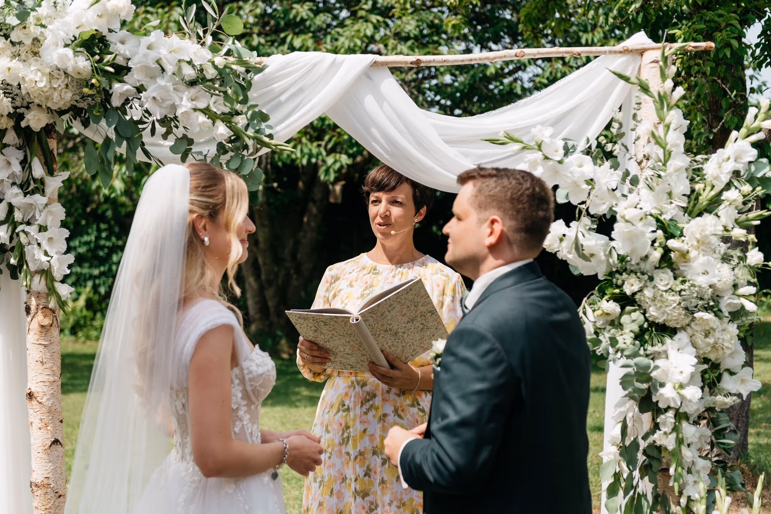 Braut und Bräutigam stehen vor Anna Fasciani - freie Traurednerin -  unter einem blumengeschmückten Bogen im Freien.