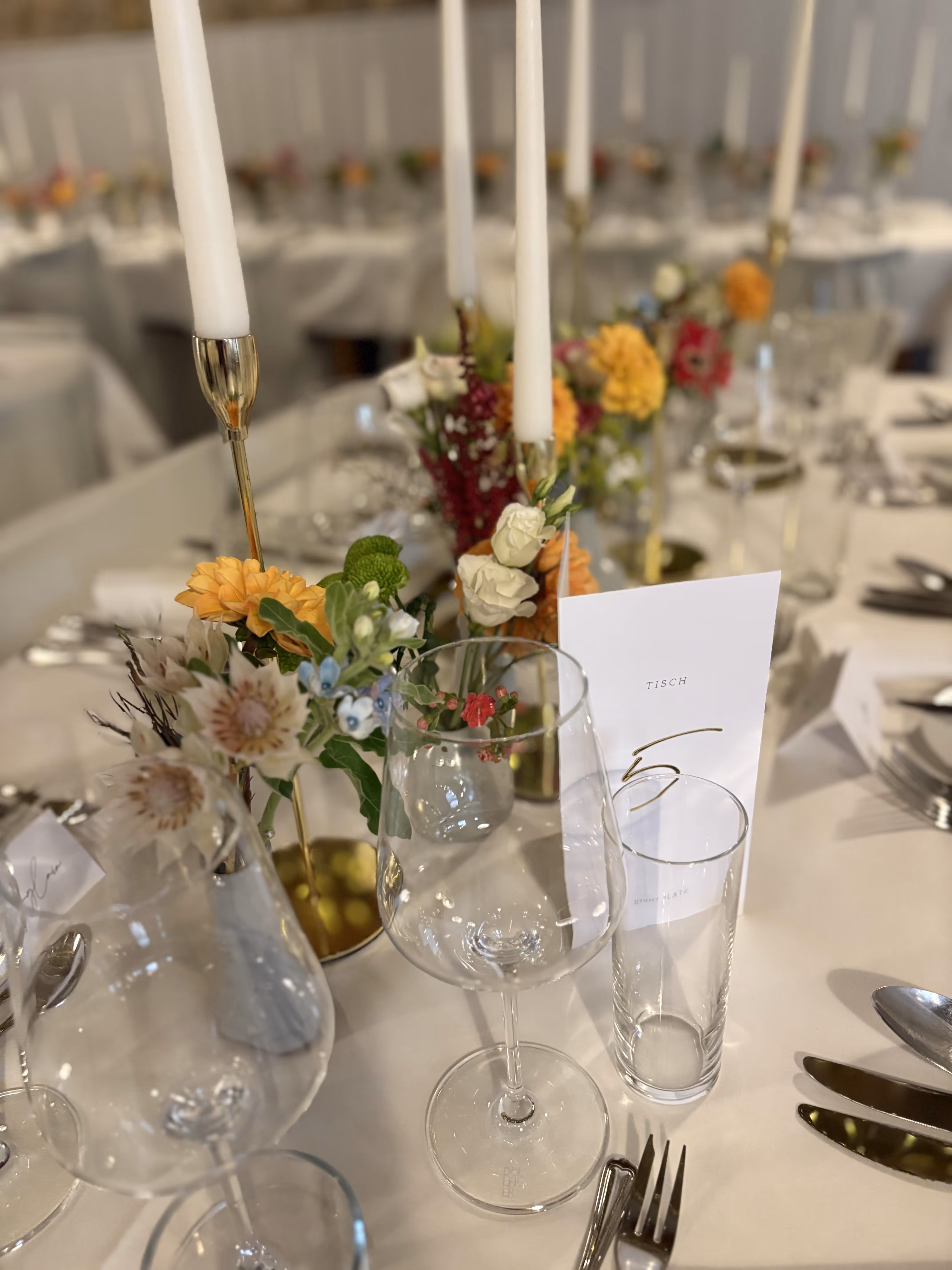 Festlich gedeckter Tisch mit Wein- und Wassergläsern, Kerzenhaltern und bunten Blumendekorationen, dazu eine Menükarte mit der Aufschrift "Tisch 5".
