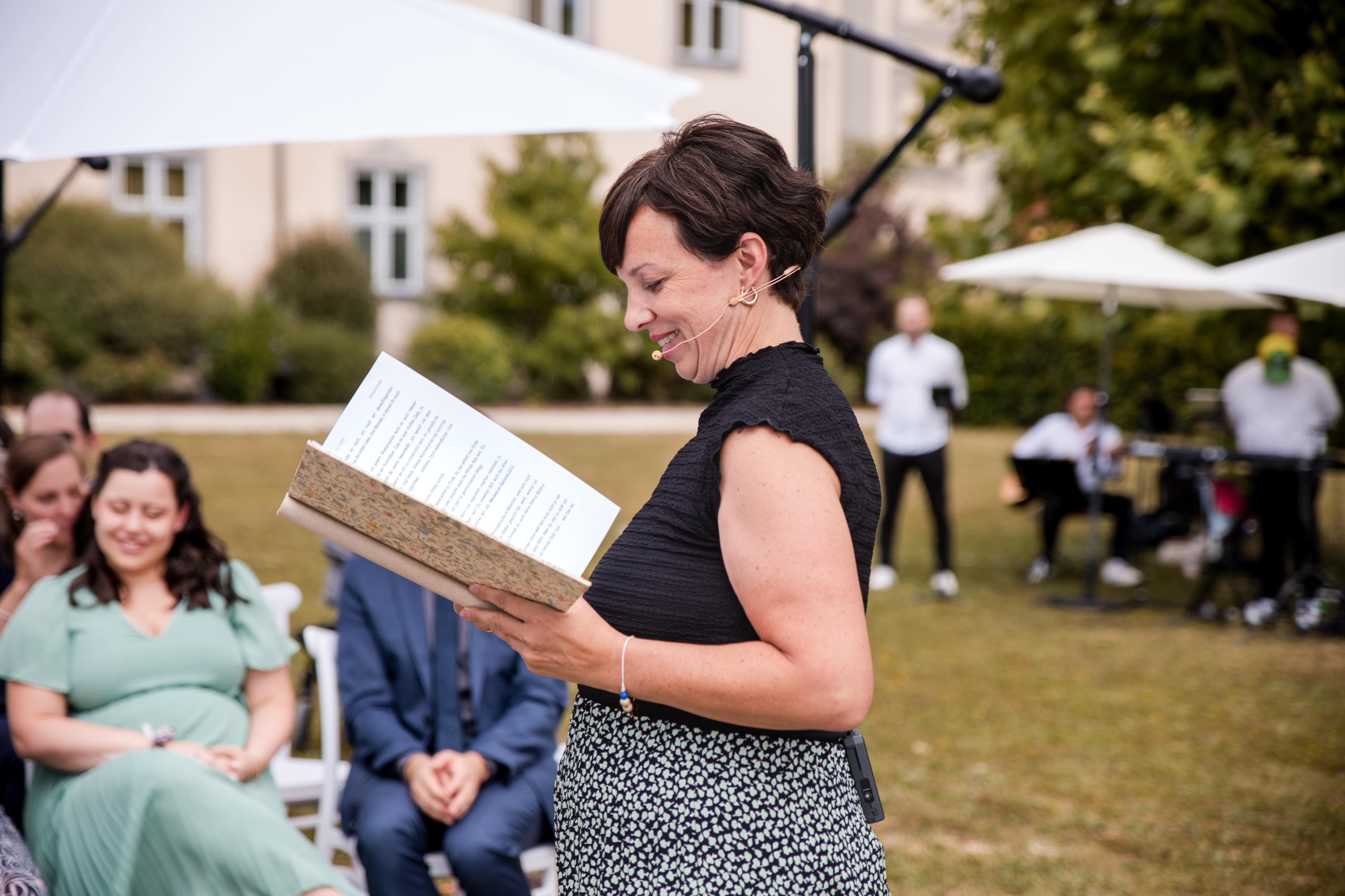 Anna Fasciani - freie Traurednerin -  liest lächelnd aus einem Buch bei einer Outdoor-Veranstaltung mit sitzendem Publikum im Hintergrund.