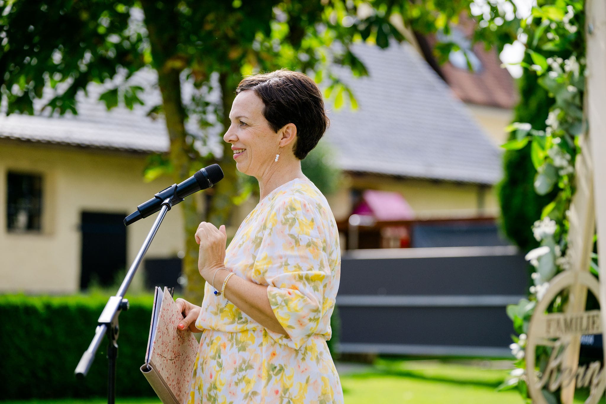 Anna Fasciani - freie Traurednerin -  in einem hellen Blumenkleid spricht bei einer Trauungszeremonie in einem Garten mit einem Mikrofon und hält ein Notizbuch.