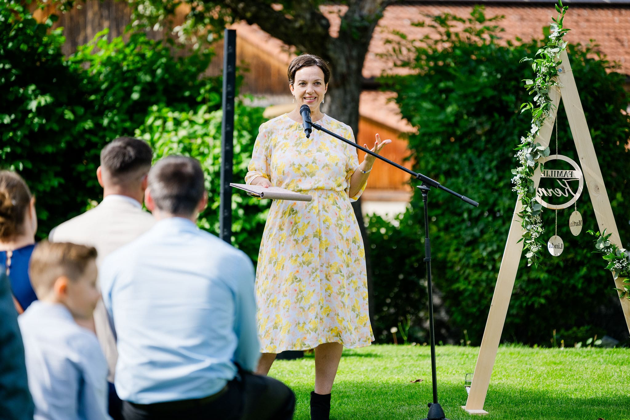 Anna Fasciani - freie Traurednerin -  in geblümtem Kleid hält eine Rede mit Mikrofon vor einer kleinen Gruppe von Menschen im Freien.