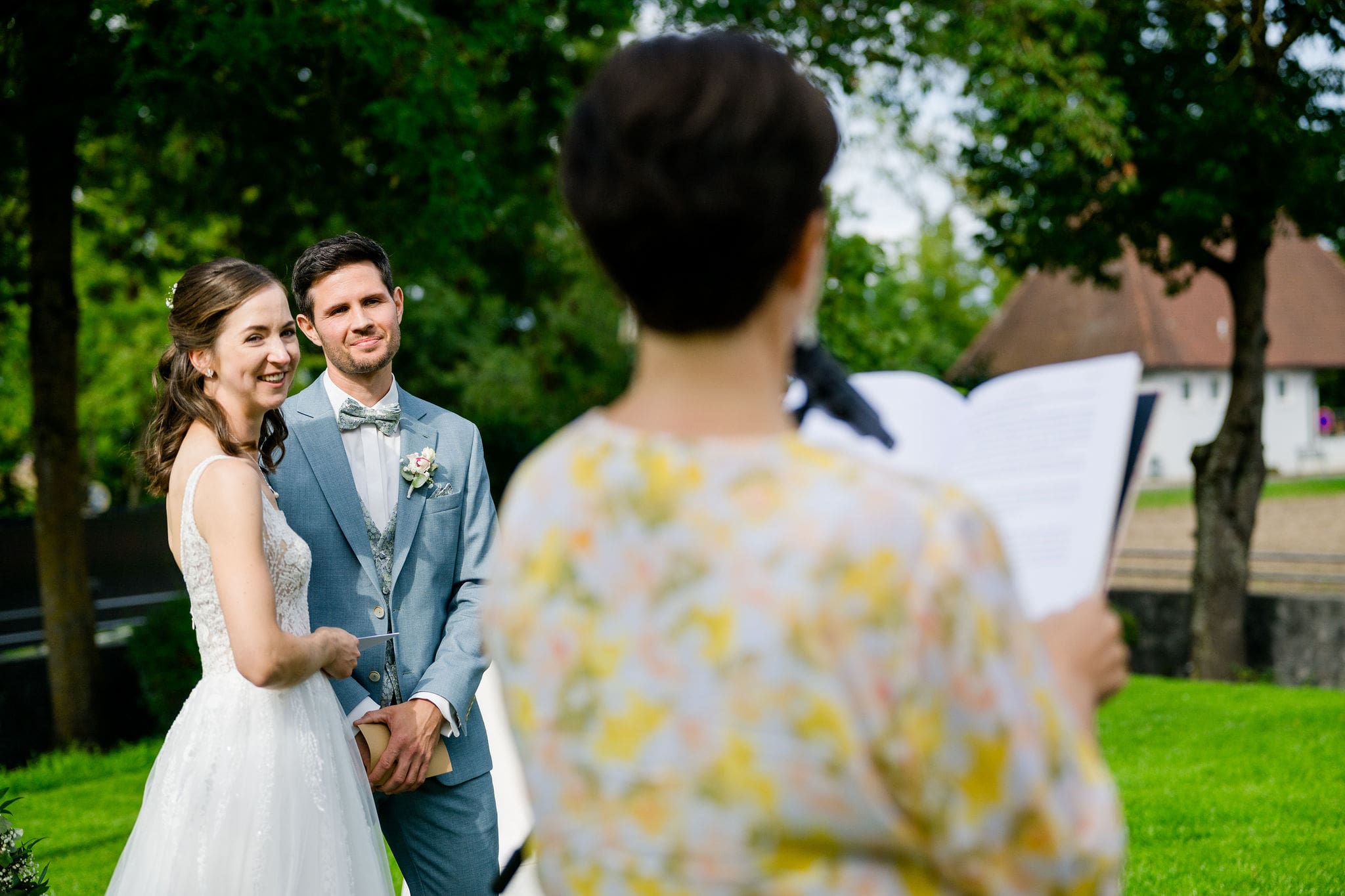 Braut und Bräutigam lächeln während der freien Trauung im Freien, mit Anna Fasciani - freie Traurednerin - , die eine Rede hält.