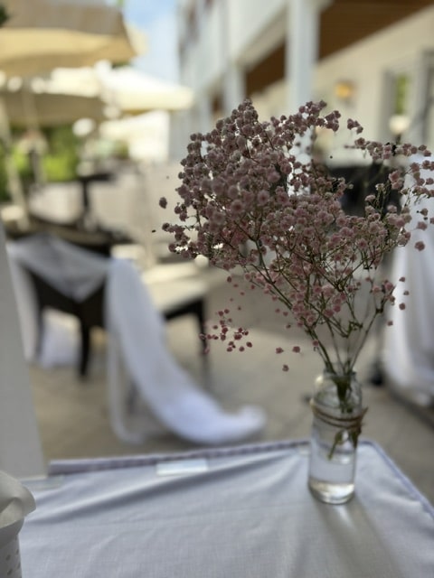 Verwischter Hintergrund mit eingedecktem Tisch und Stuhl, im Vordergrund Glasvase mit rosa Schleierkraut auf weißem Tischtuch.