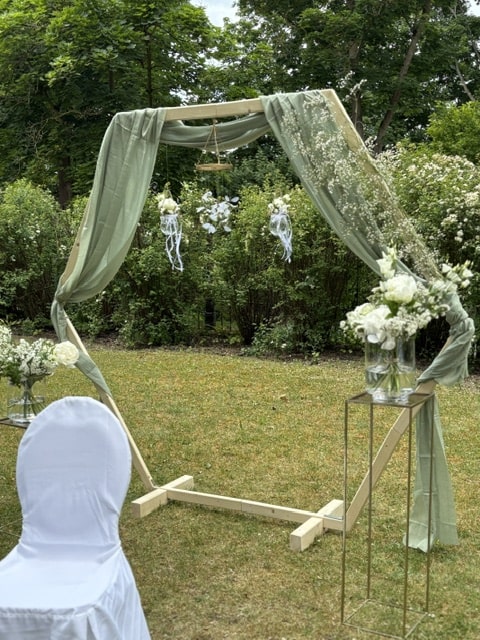 Freie Trauungszeremonie im Garten mit einem hölzernen Dekobogen, der mit grünem Tuch und weißen Blumen geschmückt ist, neben einem Stuhl mit weißem Stuhlhussen.