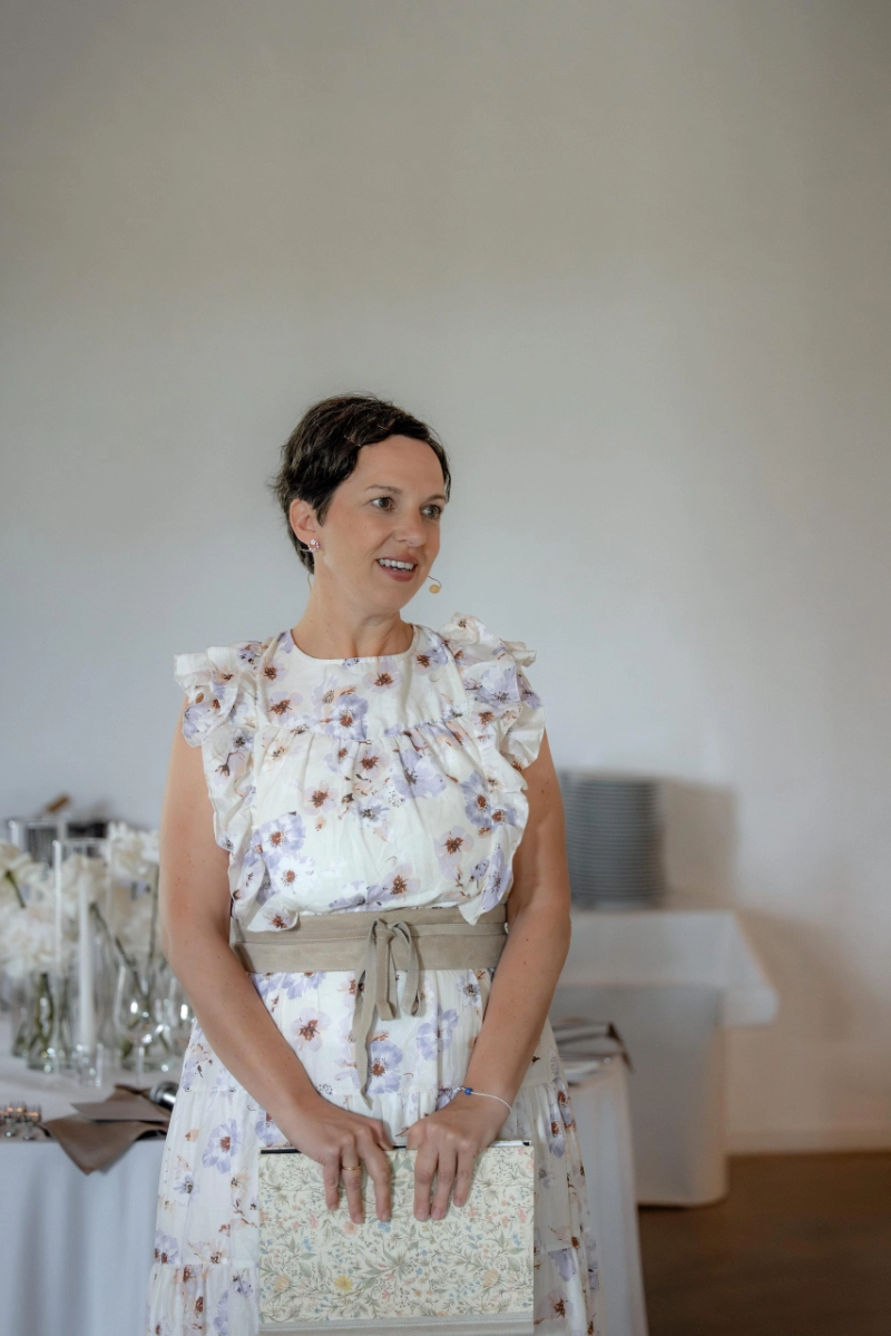 Anna Fasciani - freie Traurednerin -  in weißem Blumenkleid mit Mikrofon hält ein dekoriertes Buch bei einer Indoor-Veranstaltung.