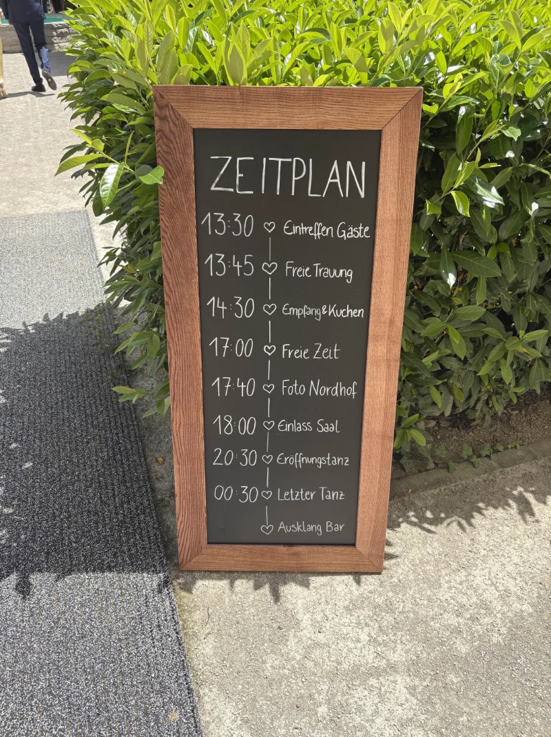 Zeitplan auf einer Tafel mit Holzrahmen vor grünen Büschen, zeigt Zeit und Aktivitäten für eine Veranstaltung von 13:30 bis 00:30 Uhr.