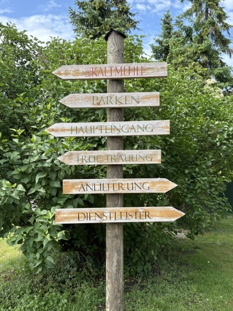 Holzscheiben-Wegweiser im Grünen mit Hinweisen zu Kaltmühle, Parken, Haupteingang, Freie Trauung, Anlieferung und Dienstleister.