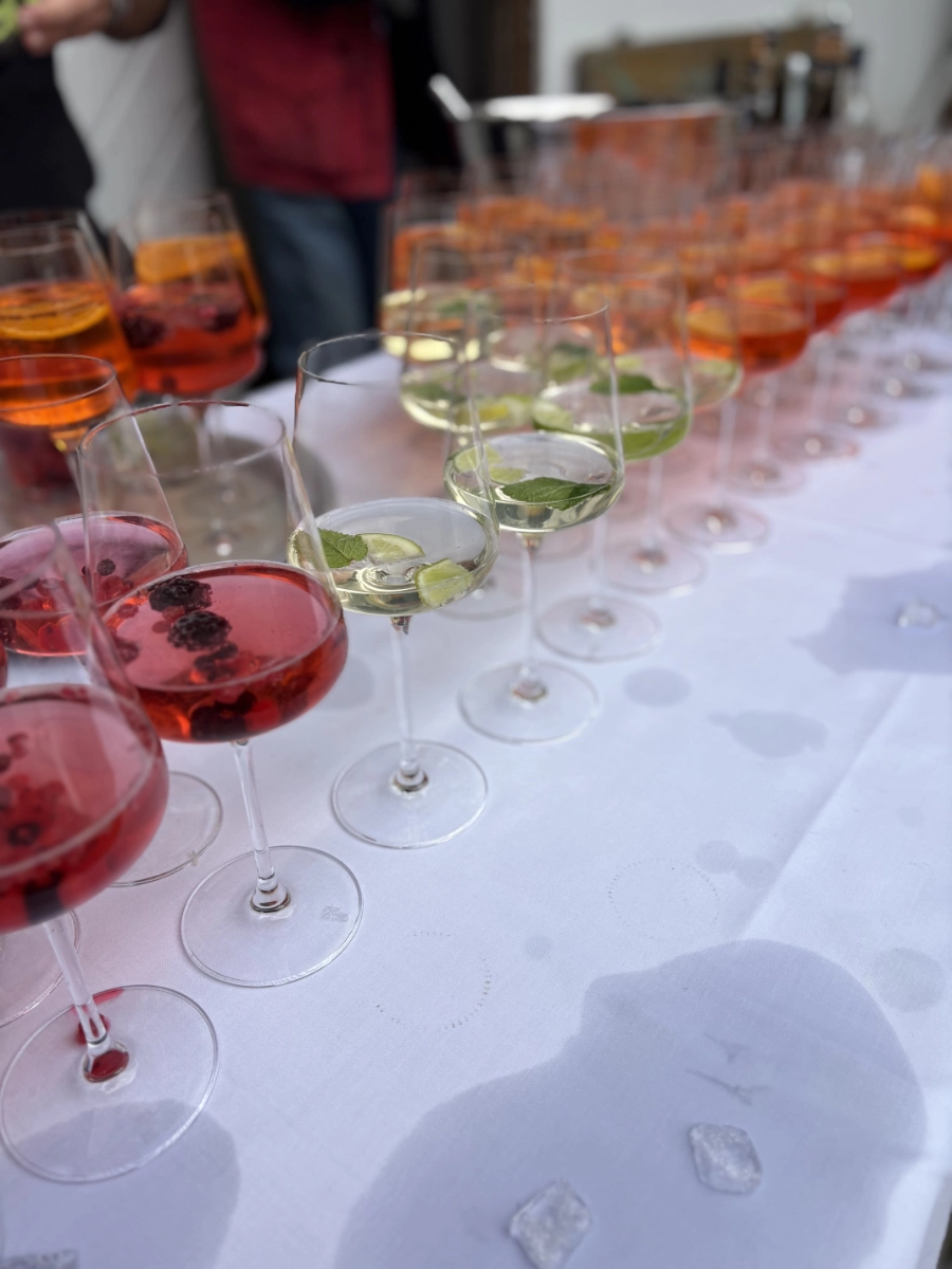 Reihe von Weingläsern mit roten, orangefarbenen und weißen Getränken auf weißem Tischdecke, einige mit Früchten und Minzblättern.