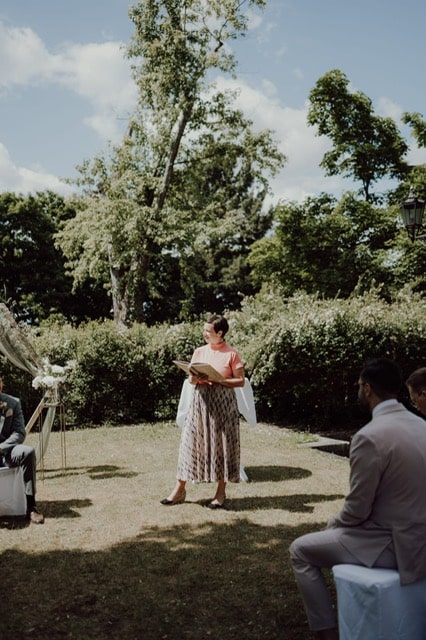 Anna Fasciani - freie Traurednerin -  steht im Freien bei einer Zeremonie, umgeben von Bäumen und Gästen, die sitzen.