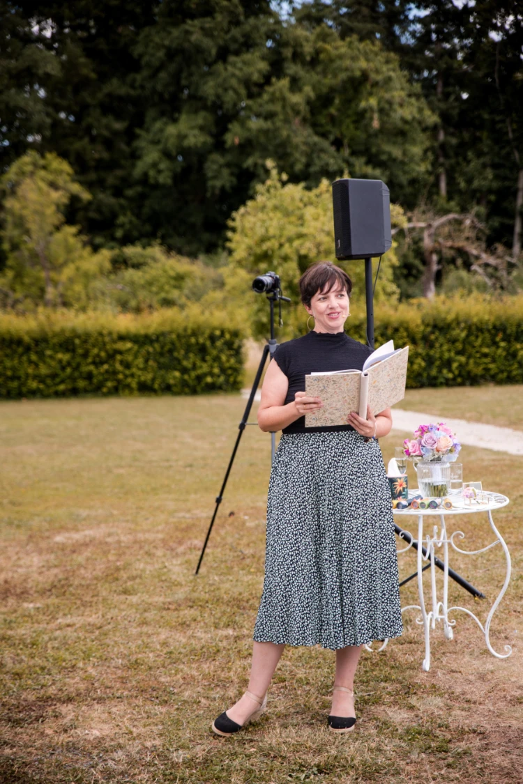 Anna Fasciani - freie Traurednerin - liest aus einem Buch im Freien neben einem weißen Tisch mit Blumen und Gläsern, hinter ihr steht eine Kamera auf einem Stativ.