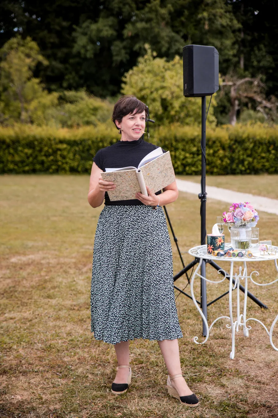 Anna Fasciani - freie Traurednerin -  mit kurzem dunklem Haar liest aus einem Buch und steht auf einer Wiese neben einem Tisch mit Blumen und Gläsern.