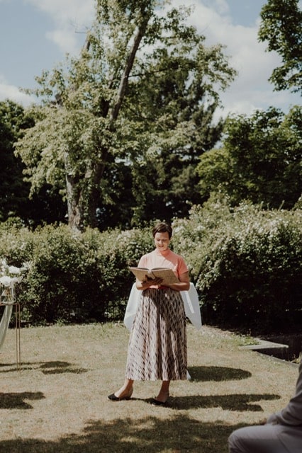 Anna Fasciani - freie Traurednerin -  steht im Freien vor Sträuchern und Bäumen und liest aus einem Buch bei sonnigem Wetter.