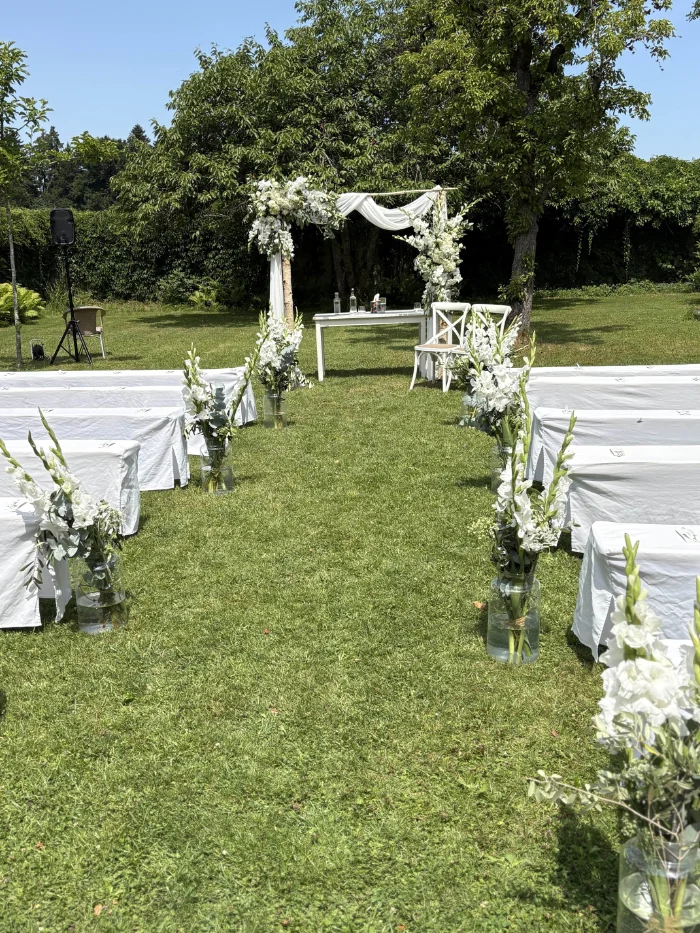 Freie Trauungszeremonie im Freien mit weißen Blumen, weißen Stühlen und Bänken auf grünem Rasen unter blauem Himmel.