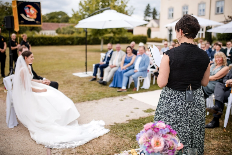 Anna Fasciani - freie Traurednerin -  hält eine freie Trauung im Freien, Brautpaar sitzt links, Gäste im Hintergrund.
