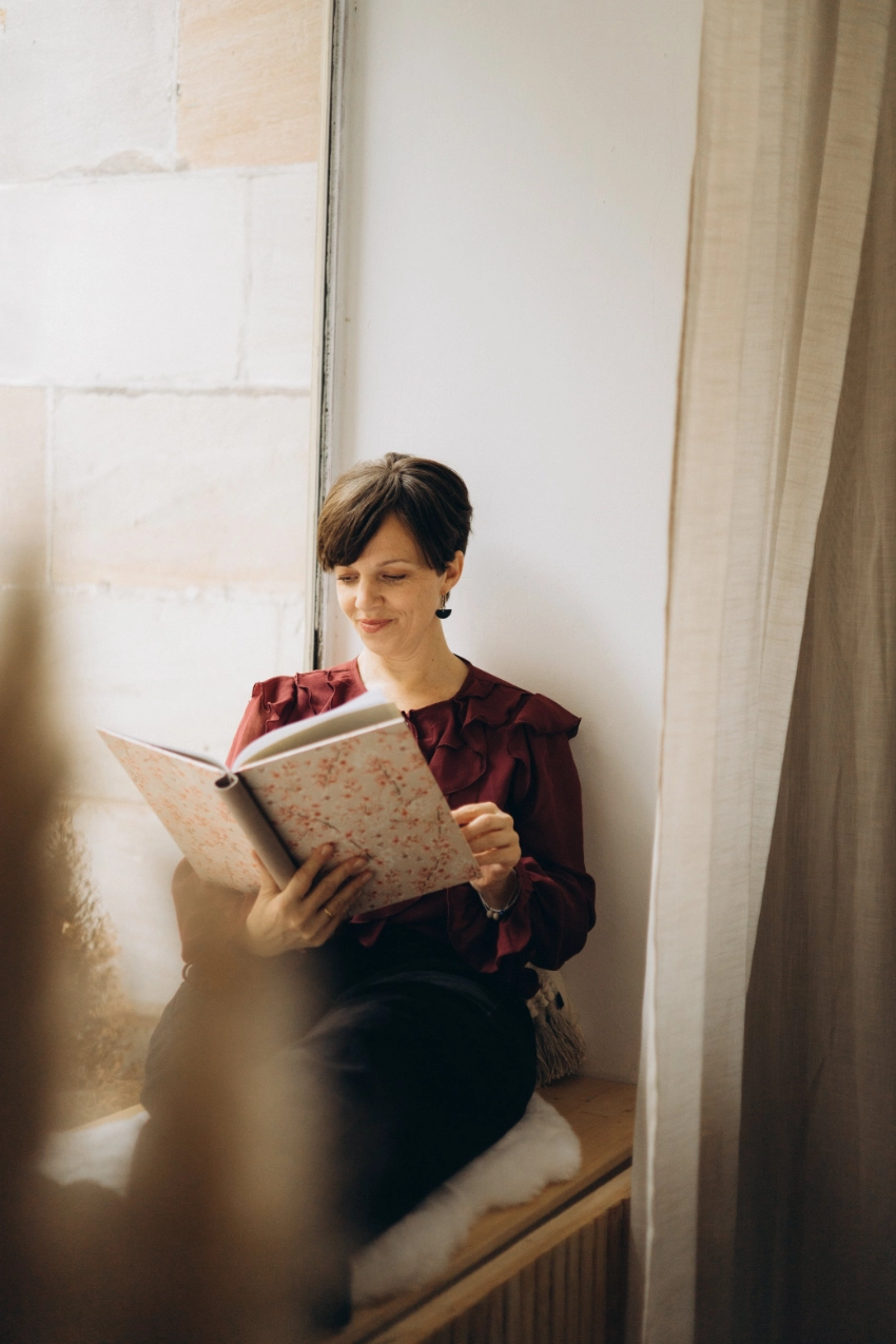 Die Freie Trauerrednerin Anna Fasciani liest lächelnd ein Buch mit floralem Umschlag am Fensterplatz.