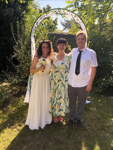 Drei lächelnde Personen stehen im Garten unter einem dekorativen Bogen mit grünen Blättern; die linke Person trägt ein weißes Kleid mit Blumenkranz und hält einen Brautstrauß.