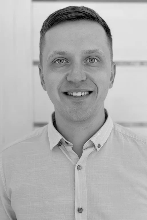 Smiling young man with short hair wearing a light button-up shirt against a blurred background.