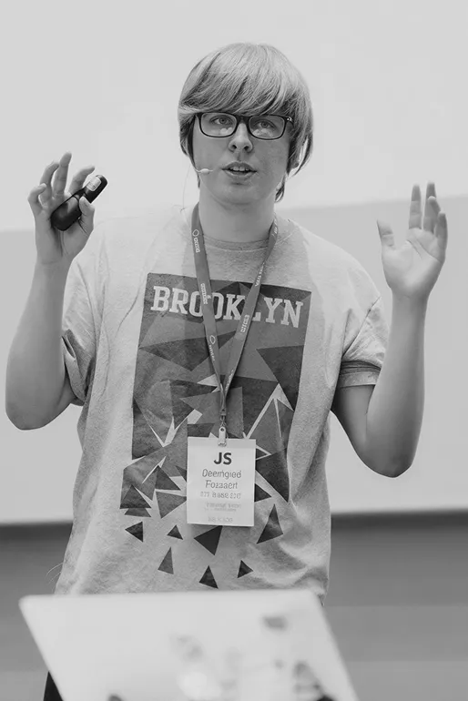 Person wearing glasses and a Brooklyn t-shirt speaking with hands raised, holding a presentation remote, wearing a conference badge labeled 'JS'.