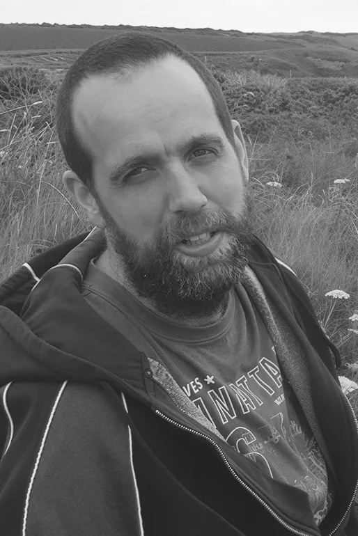 Bearded man wearing a jacket and graphic t-shirt sitting outdoors with grassy hills in the background.