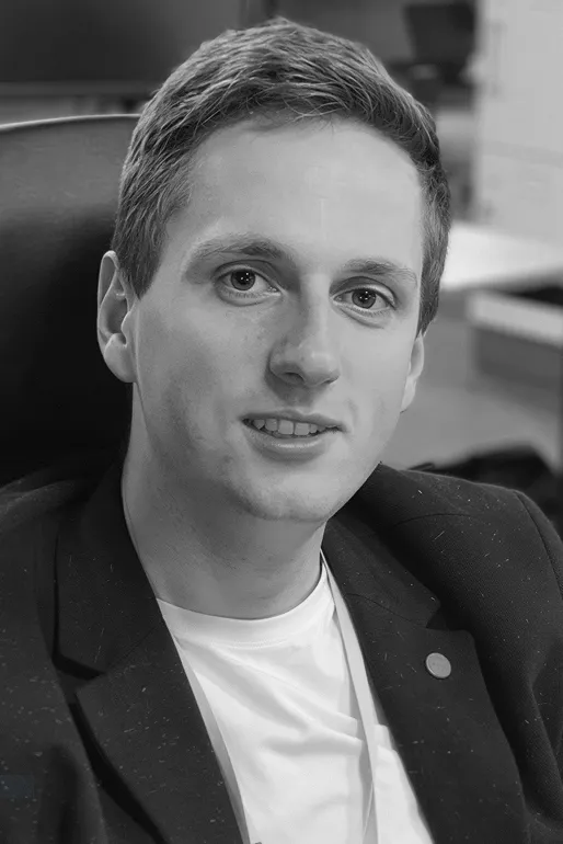 Black and white portrait of a young man with short hair wearing a blazer and white shirt, seated and looking at the camera.
