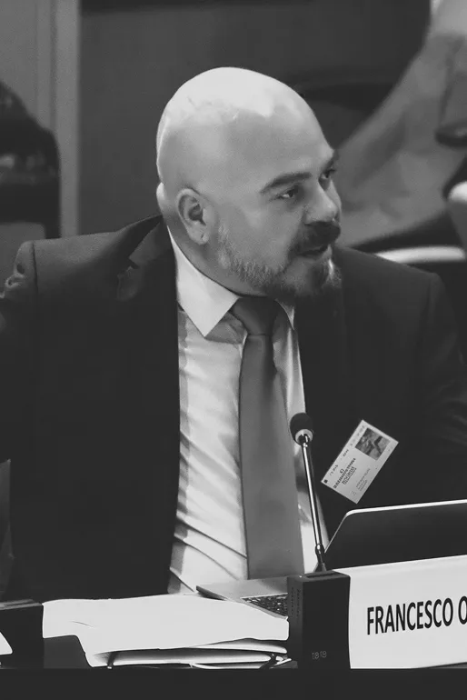 Man with bald head and beard wearing a suit and tie speaking at a conference table with a microphone and nameplate.