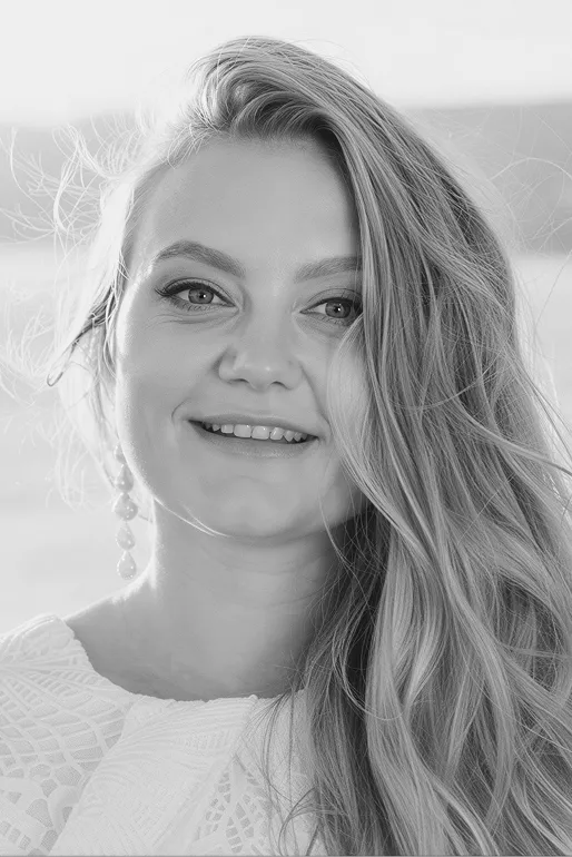 Smiling woman with long wavy hair wearing a white textured top and dangling pearl earrings.