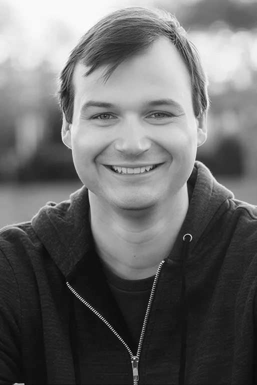 Smiling man with short hair wearing a dark zip-up hoodie, outdoors with blurred background.