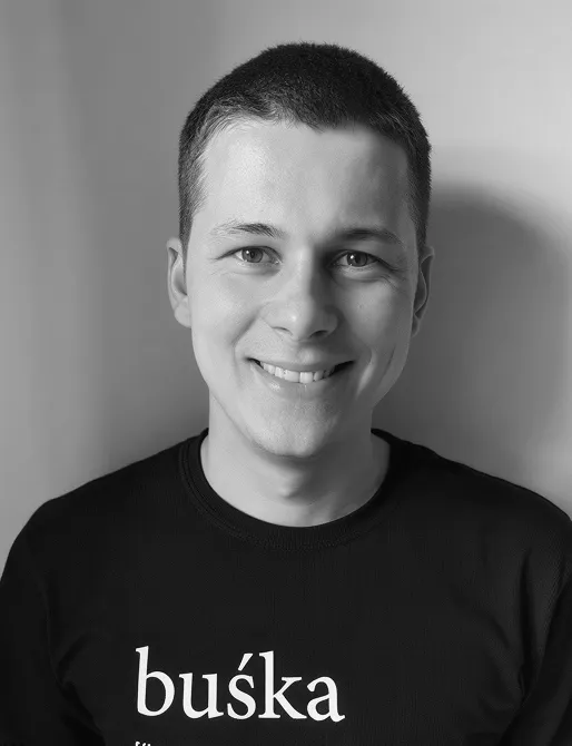 Portrait of a smiling young man with short hair wearing a black shirt with the word 'buśka' printed on it.