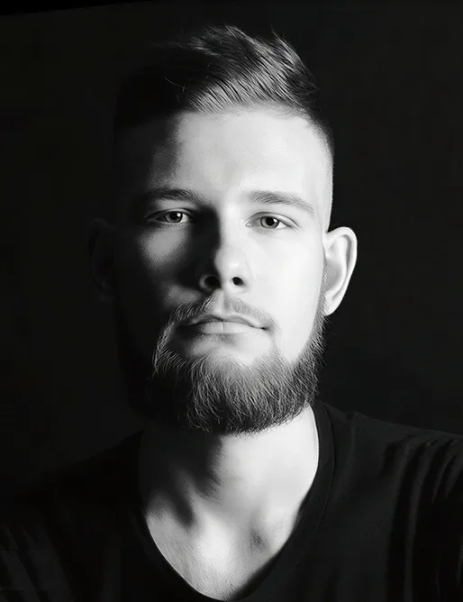 Black and white close-up portrait of a young man with a beard and short hair against a dark background.