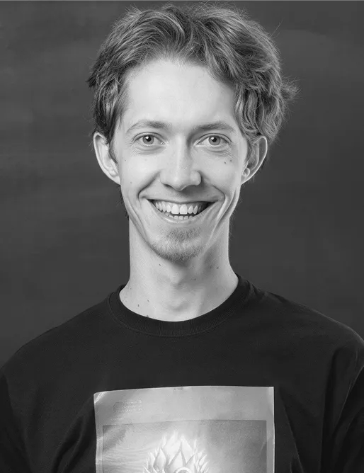 Smiling young man with light wavy hair wearing a black shirt in front of a plain dark background.
