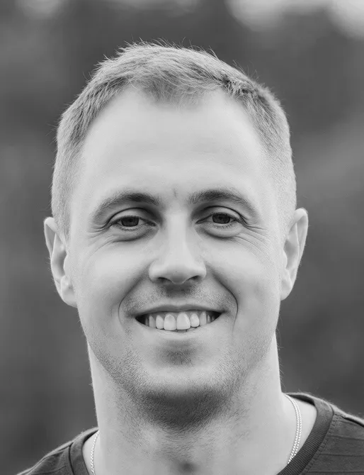 Black and white close-up portrait of a smiling man with short hair wearing a dark shirt and a thin necklace.