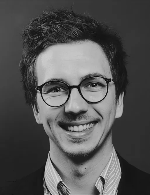Smiling young man with round glasses, tousled hair, and a striped shirt under a dark jacket against a dark background.
