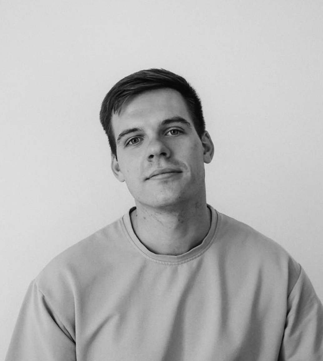 Black and white close-up portrait of a young man with short hair and a beard, looking directly at the camera with a neutral expression.