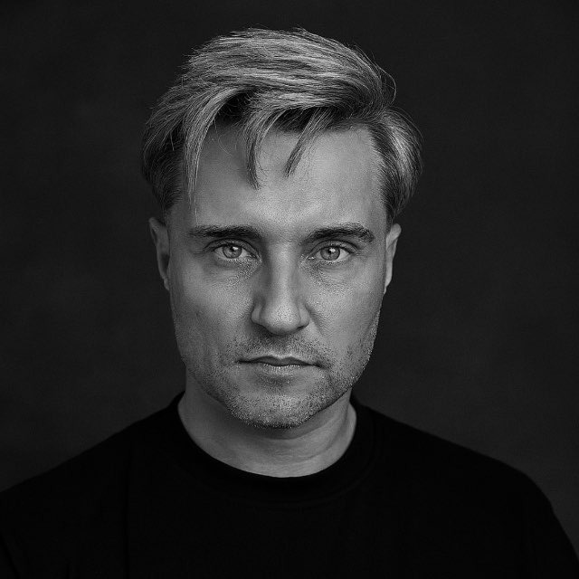 Black and white close-up portrait of a young man with short hair and a beard, looking directly at the camera with a neutral expression.
