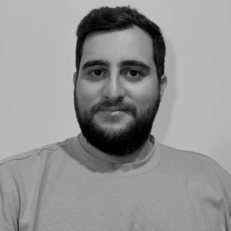Black and white close-up portrait of a young man with short hair and a beard, looking directly at the camera with a neutral expression.