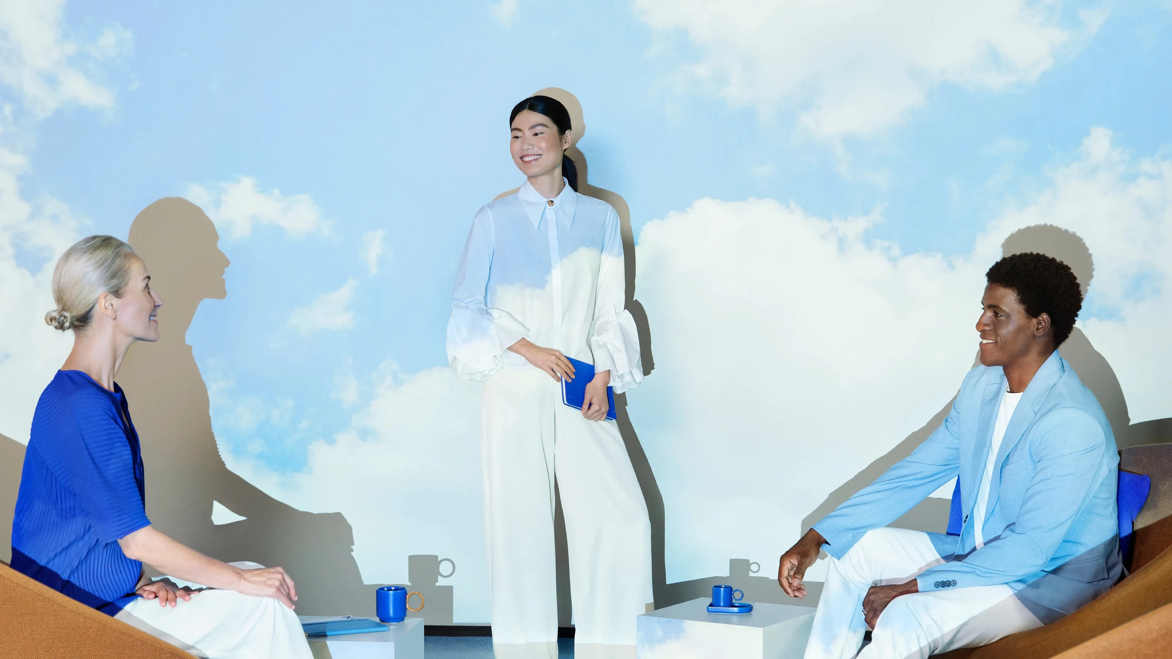 Trois personnes habillées en bleu et blanc discutent dans une pièce avec un mur projettant un ciel bleu avec des nuages.