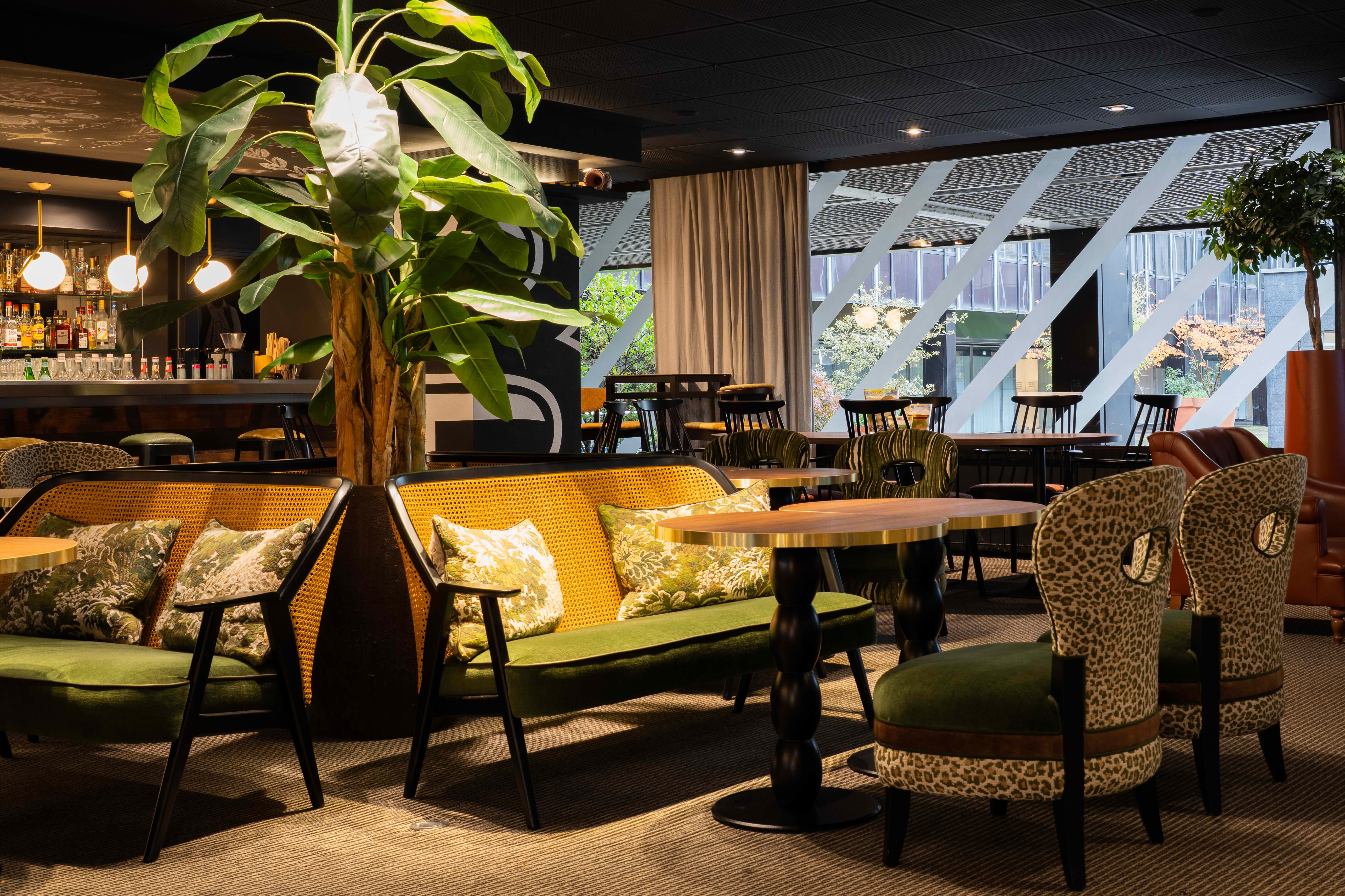 Coin salon d'un bar moderne avec canapés verts, fauteuils imprimé léopard, tables en bois, et une grande plante verte centrale.